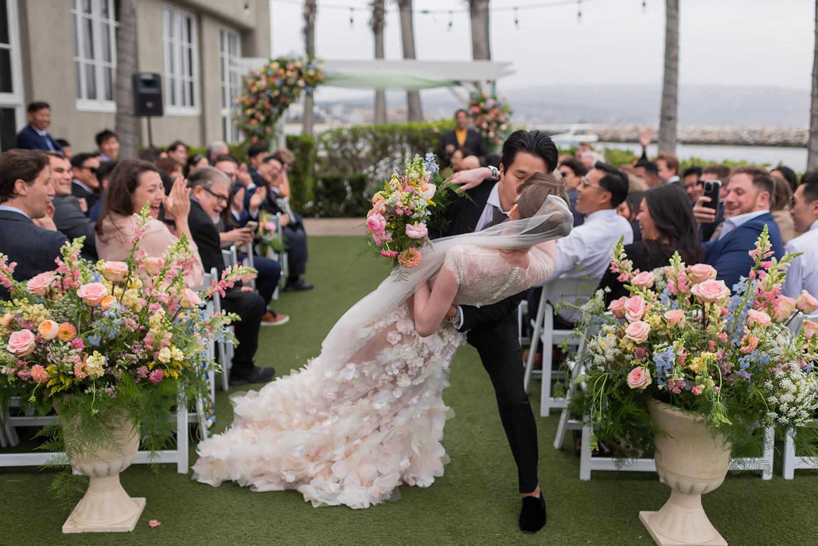 Bride and groom sharing a kiss in the aisle at an outdoor ceremony with colorful garden flowers and harbor views at The Portofino Hotel & Marina in Redondo Beach, California.