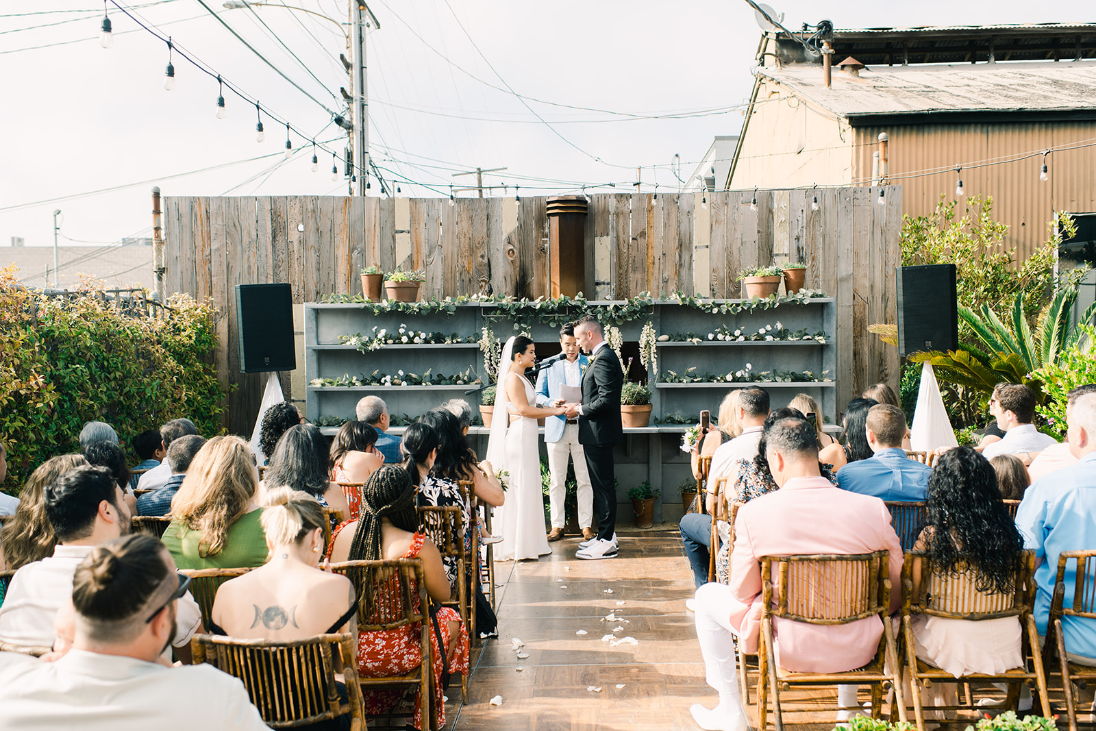 Outdoor wedding ceremony at Wwoo Kitchen in El Segundo, California, with guests seated before a wooden altar decorated with greenery and flowers.