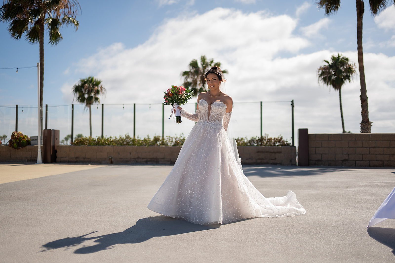 Bride in a sparkling ballgown holding a bouquet on the palm-lined oceanfront terrace at Sonesta Redondo Beach in Redondo Beach, California.