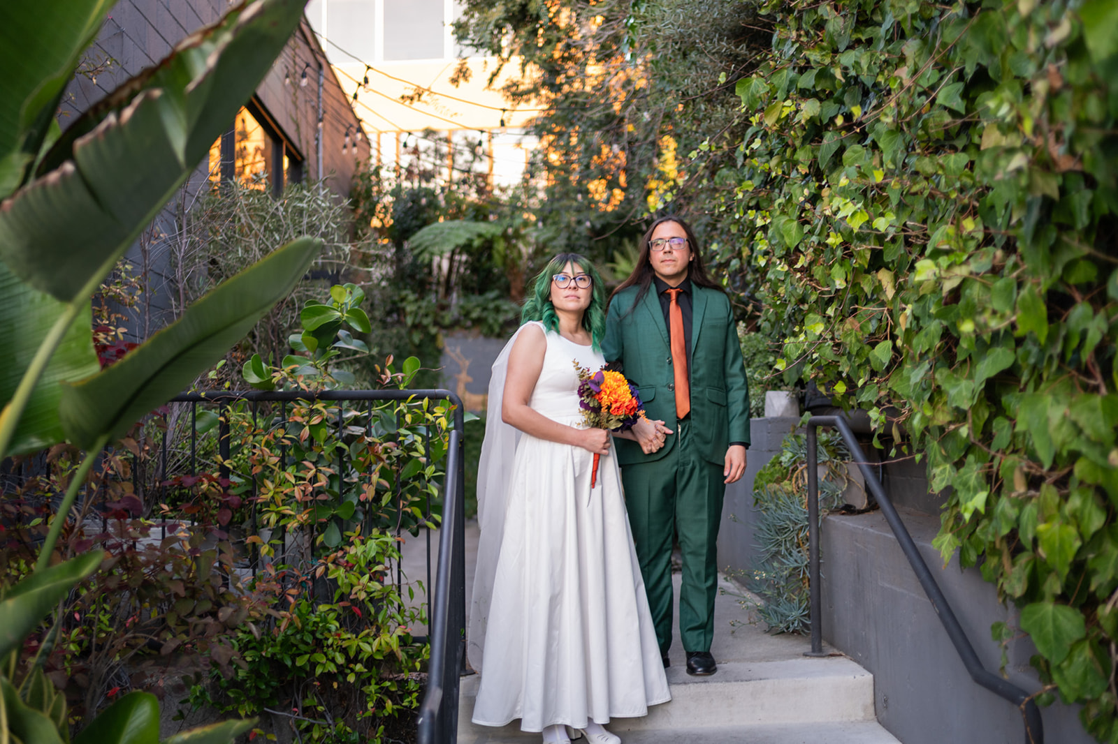 Bride and groom standing on a lush green stairway at Playa Studios in El Segundo, California, with tropical foliage and warm string lights.