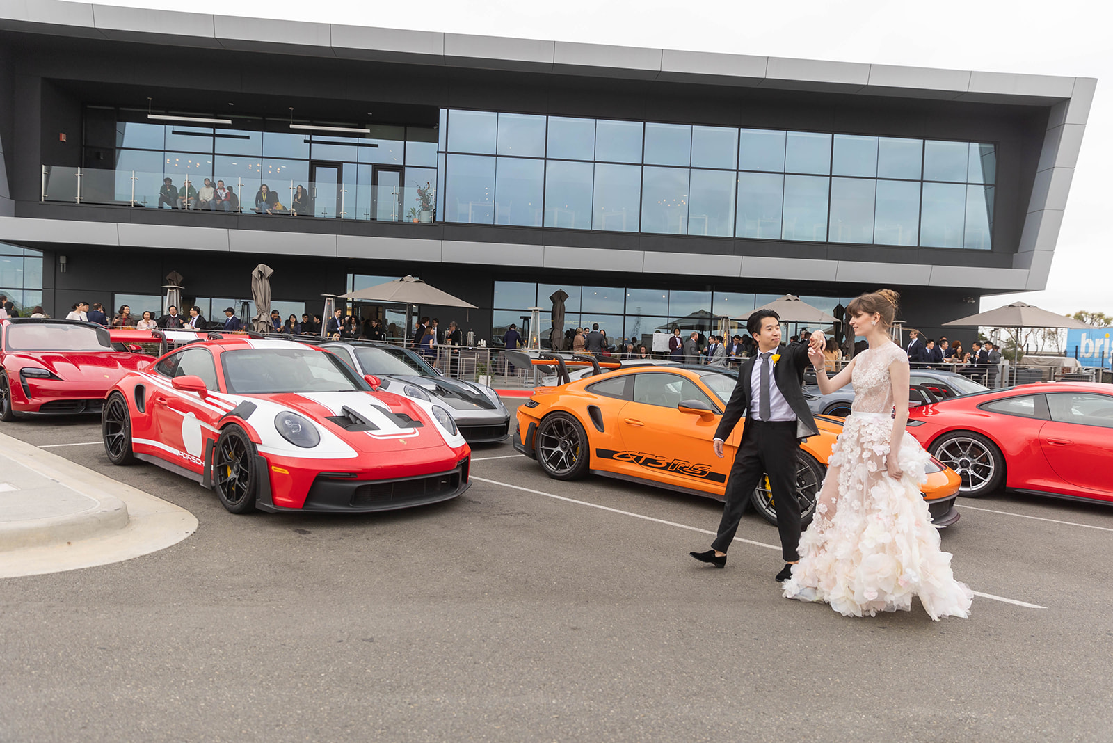 Bride and groom dancing in the Porsche Experience Center showroom in Carson, California, surrounded by colorful Porsche sports cars and modern architecture.